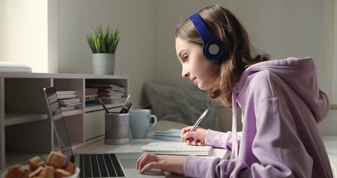 Teenage girl wearing headphones studying online from home with distance teacher. Teen school student distance learning in internet making notes, doing homework, watching video tutorial course concept.