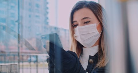 Close up portrait of young smart female volunteer in a medical mask is texting on the phone at bus stop. Quarantine COVID-19 in Europe. Coronavirus transmission in public transport.