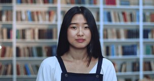 Portrait of shocked asian young female student with black hair looking at camera and covers her mouth with her palm while being very surprised in library next to book rack.
