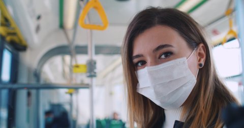 Close up portrait of smart scared female student in medical mask and gloves alone staying at public transport and looking to camera. Commute bus has few passengers.
