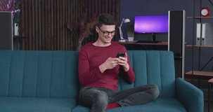 Young caucasian Man with glasses relaxing on Couch, using Smartphone in modern industrial house. Caucasian male chatting in Social Networks with Friends, feeling happy, sitting on sofa. Quarantine.