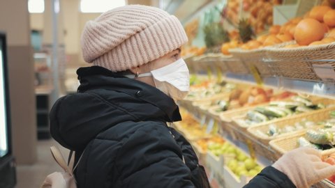 Young woman in supermarket in medical mask looks chooses and takes fruits. Coronavirus pandemic Covid-19 virus
