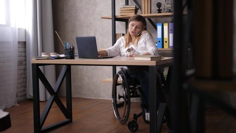 Female with disabilities holding cat pet on her knees, talking on cellphone, typing on computer while working at home. Busy woman with differing abilities during multi-task remote work in home office