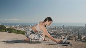 Athletic woman watching video on tablet computer during yoga training outdoor. Flexible girl doing yoga pose on city background. Focused fit woman stretching body at city view