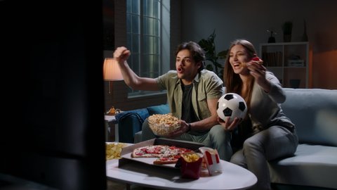 Funny young emotional caucaian sport fan couple watching a game together, eating popcorn and pizza and reacting to their team playing 4k footage