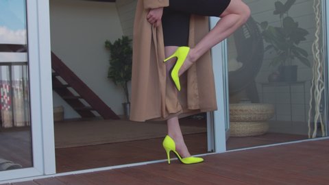 Close-up of slim elegant female legs in stylish yellow high heel shoes standing on wooden terrace. Low section of graceful woman in high heels wearing coat and leggings showing perfect slender legs.