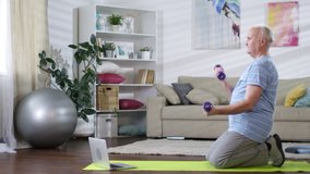 Tracking shot of senior man standing on knees on mat in the living room, watching video workout on laptop and doing dumbbell curls while training at home