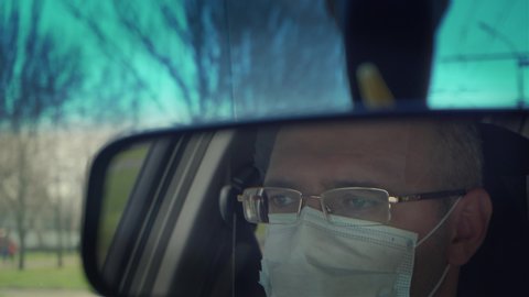 Reflection in back view mirror of man in protective medical mask driving car through city during outbreak of dangerous disease around world. Male in vehicle on town