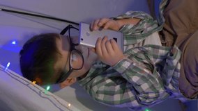 child uses phone in the evening or night sitting in a wigwam decorated with a garland at home, smartphone vertical screen