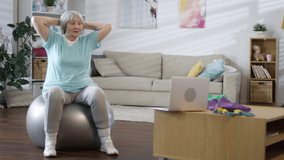 Senior woman holding hands behind head and doing fit ball seated hip rotation while following online workout on laptop at home