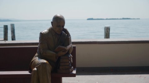 GARDONE RIVIERA, ITALY - MAY 23, 2019: wooden sculpture depicting well-known Italian poet Gabriele D'Annunzio reading a book on a bench on the promenade of Lake Garda. Sunny summer day