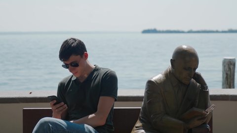 GARDONE RIVIERA, ITALY - MAY 23, 2019: young man in sunglasses with smartphone sits on bench near statue in mirror pose. Monument reads book, man reads smartphone. Funny frame