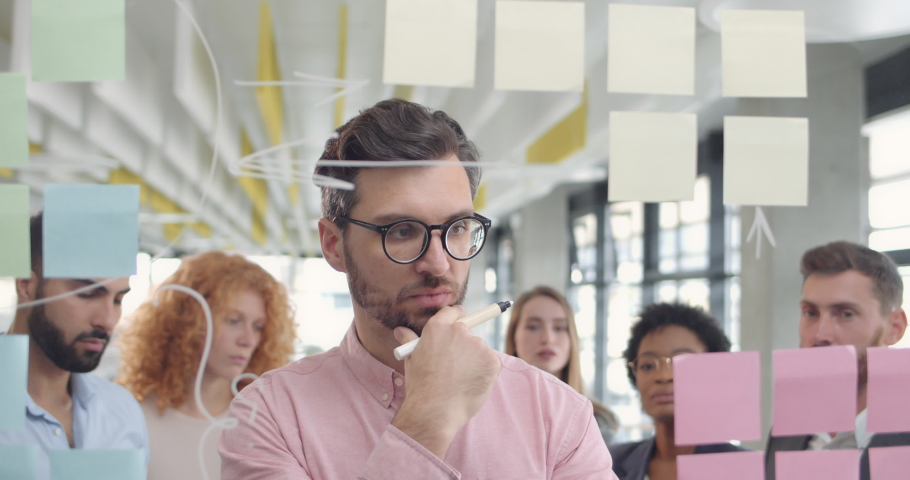 Male office worker in glases thinking and showing his colleagues new problem solution. Handsome team leader sharing his idea with office teammates and using glass board. Concept of creativity.