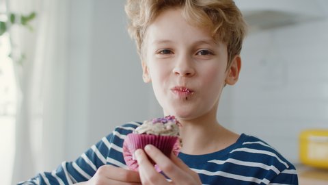 In the Kitchen: Adorable Boy Eats Creamy Cupcake with Frosting and Sprinkled Funfetti. Cute Hungry Sweet Tooth Child Bites into Muffin with Sugary Frosting