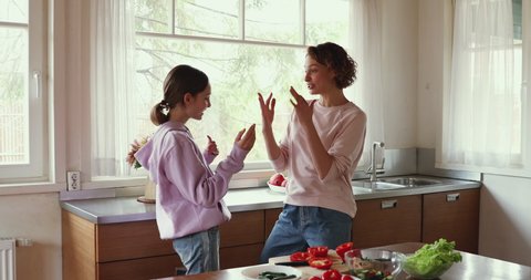 Young happy family mom and teen daughter enjoy funny dance in kitchen cooking together. Cheerful adult parent mother having fun with adolescent teenage girl preparing healthy salad at home together.