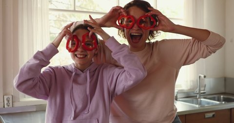Funny young mom and teenage daughter making pepper glasses having fun. Happy teen girl helping mum in kitchen laughing, looking at camera. Cheerful vegan family enjoying cooking activity, portrait.