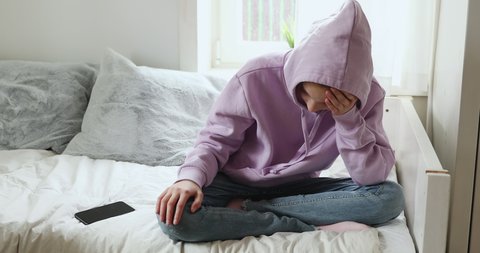 Depressed teenage girl sitting alone at home with smart phone. Upset adolescent teenager outcast wearing hood feeling worried or scared about social media cyber bullying mobile message concept.