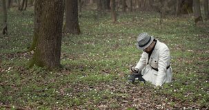 Confident man in trendy outfit sitting on ground at forest and looking through magnifying glass on small white flowers. Concept of adventure and science