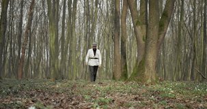 Full length portrait of bearded man in black trousers, white jacket and hat walking among green forest with walking stick. Mature botanist spending time on fresh air.