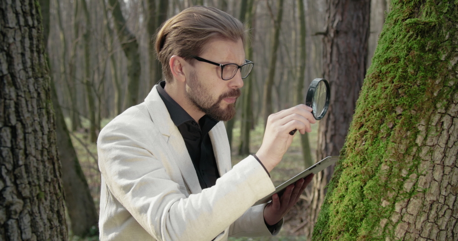 Confident bearded man in eyewear, black shirt and white jacket standing at forest with magnifying glass and examining green moss. Concept of technology and discovering