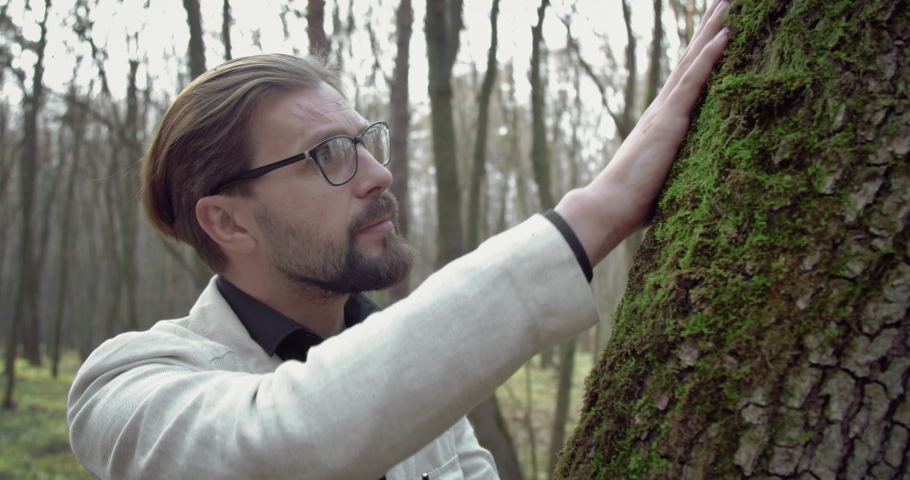 Confident bearded man in white jacket and black shirt carefully touching green moss on old tree. Mature botanist in eyeglasses enjoying favorite hobby outdoors.