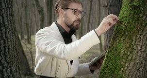 Competent botanist in eyewear and formal outfit standing near old tree and examining green moss. Bearded man filling all information about plants in digital tablet.
