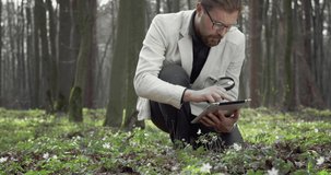 Mature botanist in white jacket and eyeglasses sitting at forest and discovering green plants with magnifying glass and digital tablet. Concept of adventure and science.
