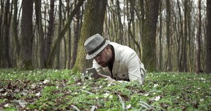 Confident botanist in white jacket and hat lying on ground at forest and taking photo of green plants on smartphone. Bearded man examining beautiful nature around.