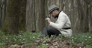 Side view of bearded man in stylish outfit and hat carefully investigating plants at forest. Mature botanist discovering green nature with loupe.