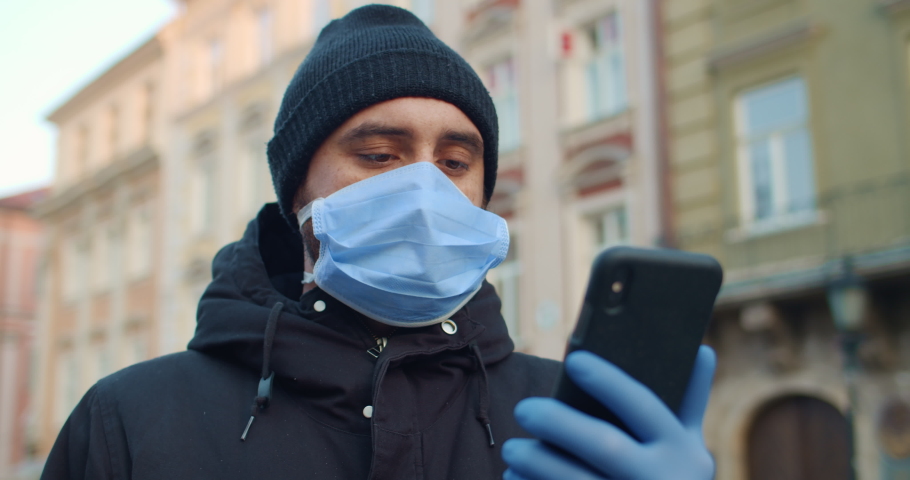 Close up view of man in protective mask and gloves looking at phone screen. Guy with medical mask a on his face standing alone and holding smartphone. Concept of coronavirus pandemic