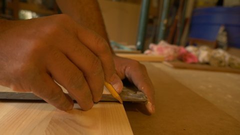 Carpenter holds a wooden plank on the work table while drawing a line with a pencil on the plank with the help of a square in his carpentry workshop.