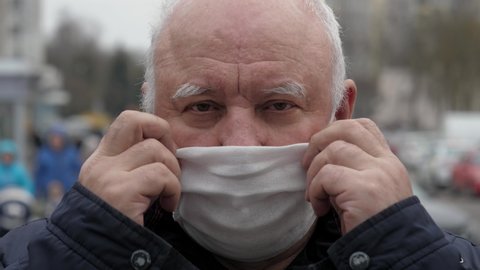 Close up portrait elderly man puts a medical mask on his face to protecting yourself from coronavirus pandemic. Standing on street, people are moving in background. Protection health and safety life.