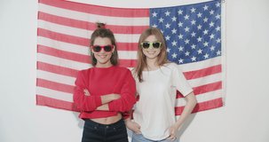 Two pretty stylish girls friends sisters celebrating independence day and having fun over national usa flag on white wall indoors. 4k video footage slow motion