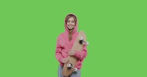 Young positive woman having fun isolated on green screen background at studio . Blonde girl dressed in pink hoodie holding skateboard. 4k raw footage video side view