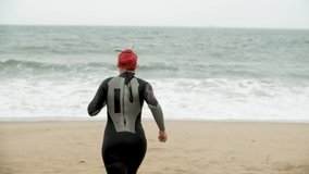 Female swimmer running on beach. Back view of young woman in wetsuit running on sand to sea waves. Swimming concept