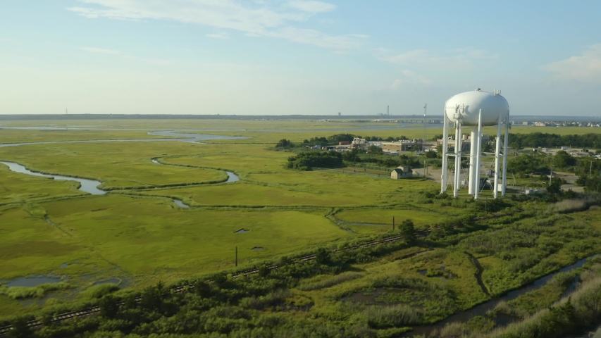 Aerial, tall white water tank tower in green swampland, sunny day