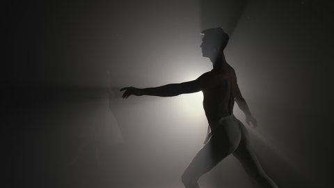 Professional choreographer is working on creating performance. Young handsome male dancer practicing in classical ballet in studio with smoke back light. Man in white tights.