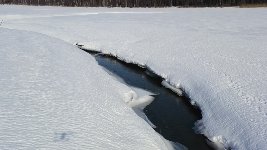 Flying in winter over a swamp covered with forest. A narrow channel of dark open water cuts through a vast expanse of snow-covered ice, winding across the frozen wetland toward a distant treeline of bare deciduous trees. The aerial perspective reveals the stark contrast between the deep black water and the bright white snow, with the channel
