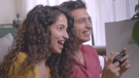 A young happy attractive married couple holding joysticks in their hands and playing an online video game together in the house. A smiling laughing man and woman spending time with each other indoors