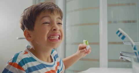 Adorable boy in pyjamas brushing his teeth thoroughly, looking in the mirror and wrinkling his face. Close-up side shot.