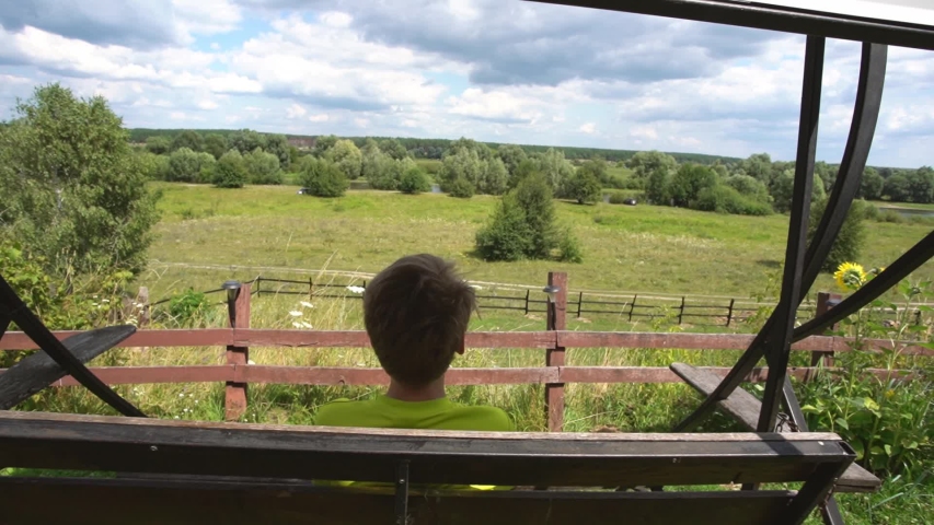 White young happy kid enjoying summer vacations in countryside sitting on wooden swing in beautiful sunny green landscape.