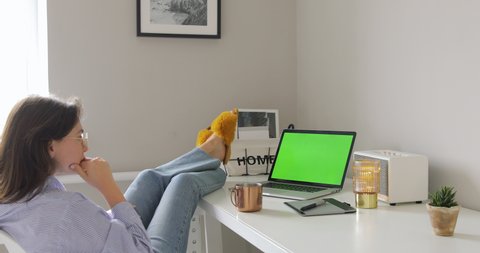 Beautiful young woman watching laptop with green screen, drinking coffee while sitting on the chair in the living room. Feet on the desk. Self isolation, Home quarantine, pandemic people, covid-19