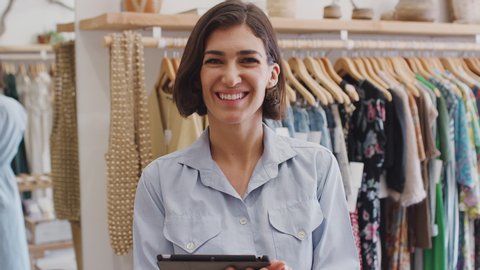 Portrait of female owner of fashion store using digital tablet to check stock on rails in clothing store - shot in slow motion