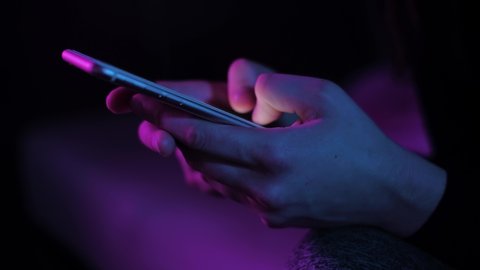 Young woman texting chatting on smart phone at night; girls finger tapping keypad, writing message on mobile devices touchscreen, blue low light; close up top side view