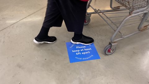 An elderly woman pushes her shopping cart over a sign on the floor of a store that warns shoppers stay 6 feet apart to comply with social distancing requirements.  	