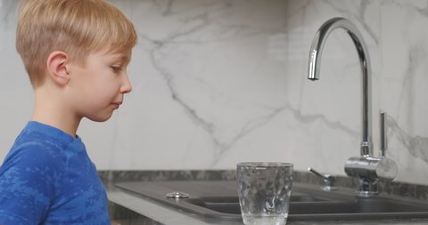 Child takes a clear glass, fills it with fresh water and drinks it. Blond boy goes to the kitchen tap opens it pours water and drinks.  