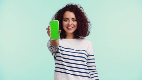 happy, curly girl showing smartphone with green screen isolated on turquoise