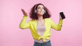 cheerful girl dancing and singing in wireless headphones isolated on pink