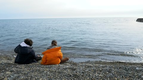 boy and girl teenagers sitting on the beach fall, looking at the sea.