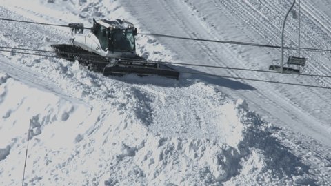 Closeup of a ski area snowplow pushing snow to build a half pipe, with a snow gun shooting man-made snow in the background.